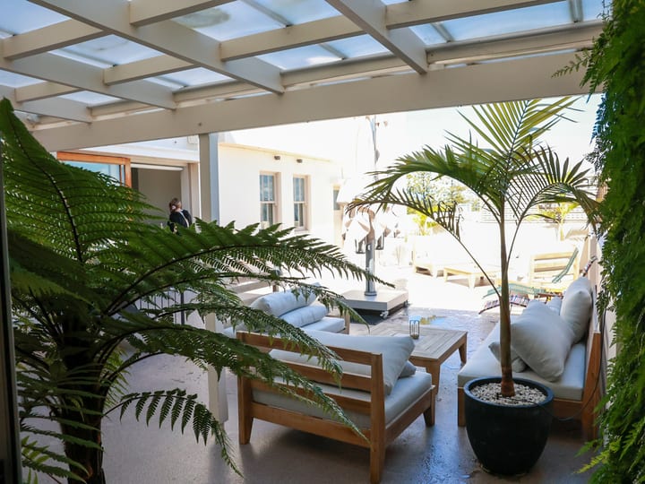 Covered outdoor terrace with white lounge furniture, potted palms, and fern wall, pergola roof
