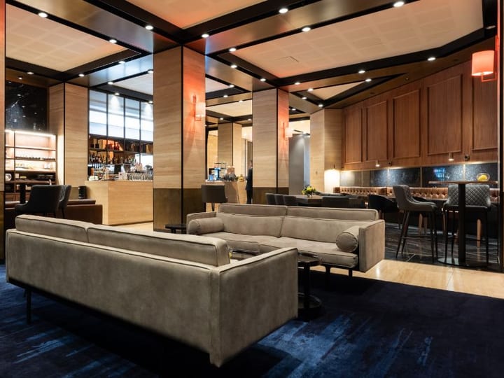 Lounge area with grey sofas, wood and mirror wall panels, warm ceiling lights, bar visible through opening