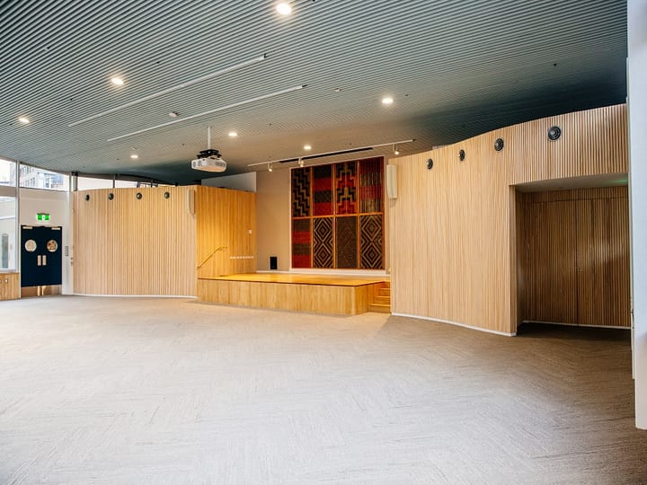 Large open hall with timber slatted walls, raised platform, tukutuku panel artwork and dark ceiling