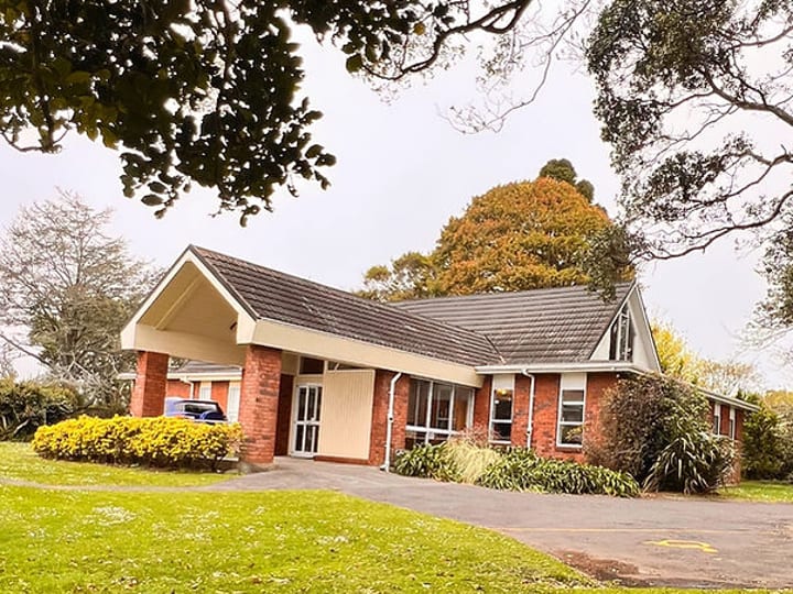 Low-set brick building with a pitched roof and covered entry, surrounded by lawn and trees