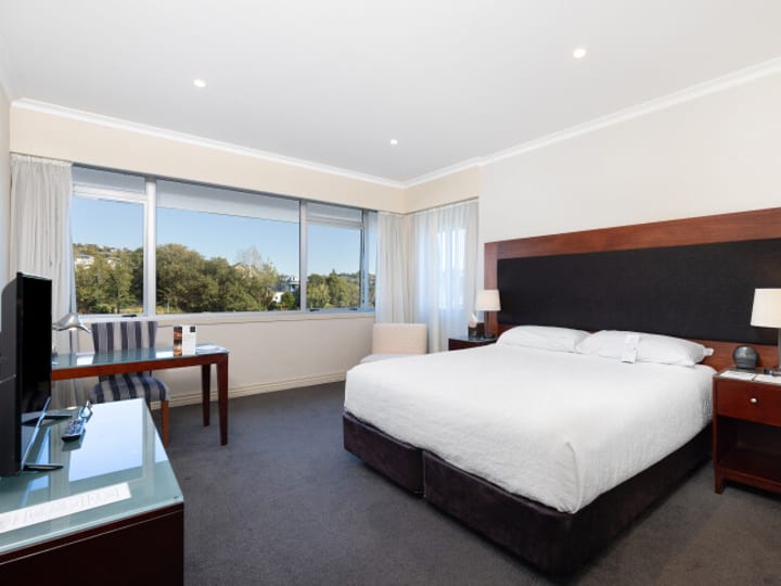Hotel room with king bed, dark timber headboard, desk and windows to green view