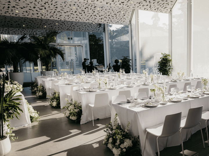 Glass-walled function room with long white tables and floral arrangements set for dining