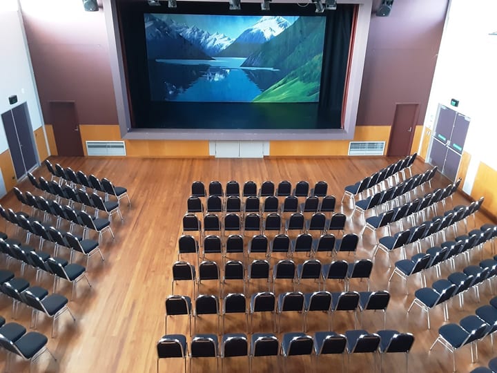 Auditorium with rows of black chairs facing a large projection screen on a raised platform, timber floor