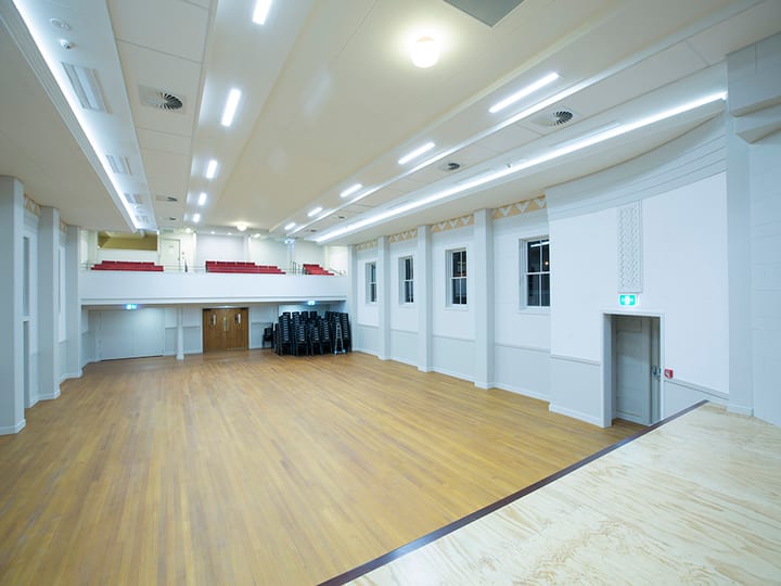 Large open hall with timber floor, balcony with red seating, and stage at far end