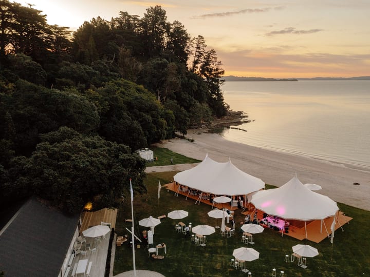 Two white peaked marquee tents on lawn beside beach at sunset with umbrellas and outdoor tables