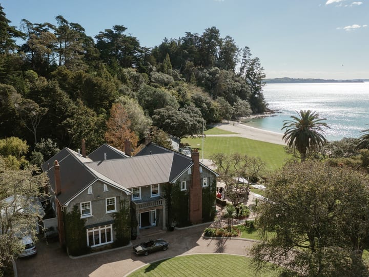 Aerial view of large historic stone manor with lawn, trees and beach frontage