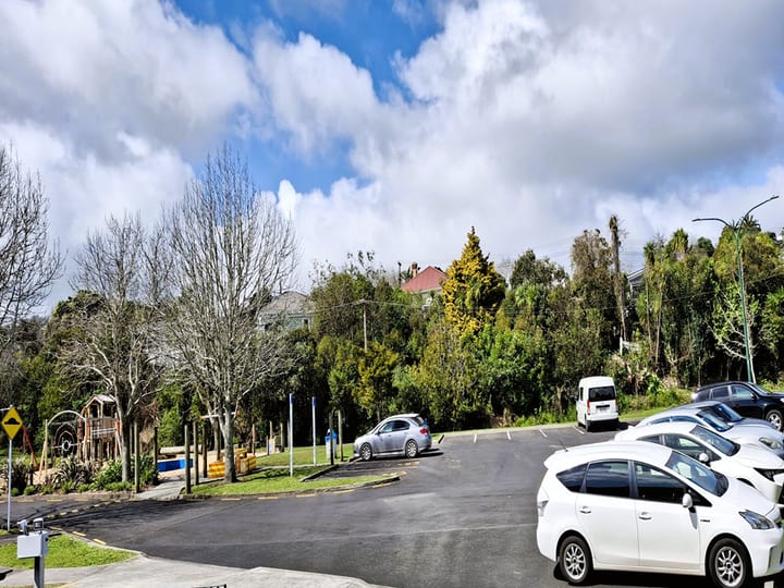 Car park area with playground and trees in background under partly cloudy sky