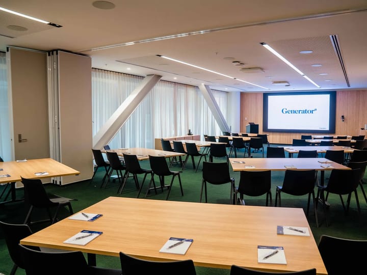 Large seminar room with rows of timber tables, black chairs, projection screen, and glazed louvre wall to one side