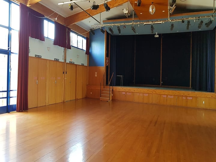 Hall with timber ceiling, stage with blue curtain, wooden floor and red curtains along windows