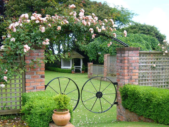 Brick garden gateway with climbing roses and black iron wagon wheels, gazebo beyond on lawn