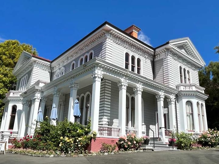White two-storey Victorian villa with ornate columns, bay windows, and flowering garden beds