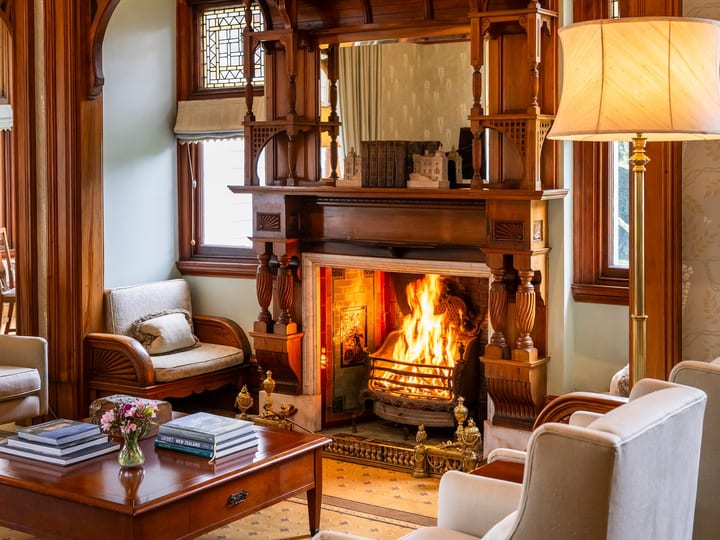 Victorian sitting room with ornate carved timber fireplace surround, lit fire, armchairs, and side tables