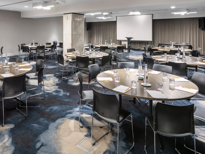 Hotel conference room with round tables, chairs, projector screen, and patterned carpet