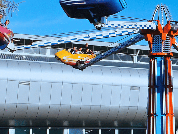 Large curved glass arena building with fairground ride in foreground