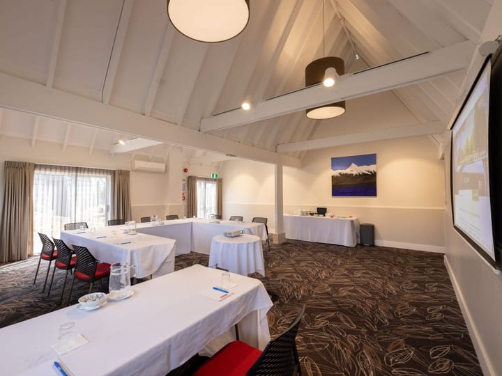 White-plastered function room with vaulted timber-truss ceiling, white tablecloths, projection screen, and patterned carpet