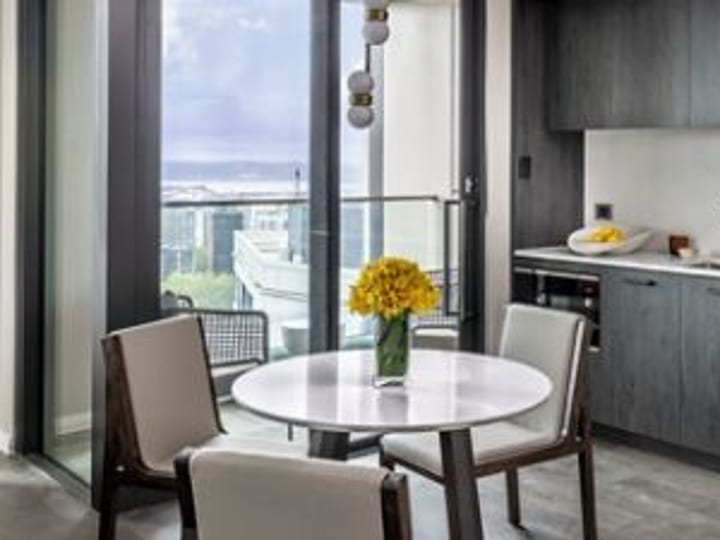 Compact suite dining area with round white table, two chairs, dark timber kitchen bench, glass balcony door