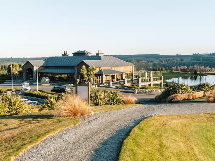 Rural resort building with metal roof beside a pond, viewed from a gravel driveway