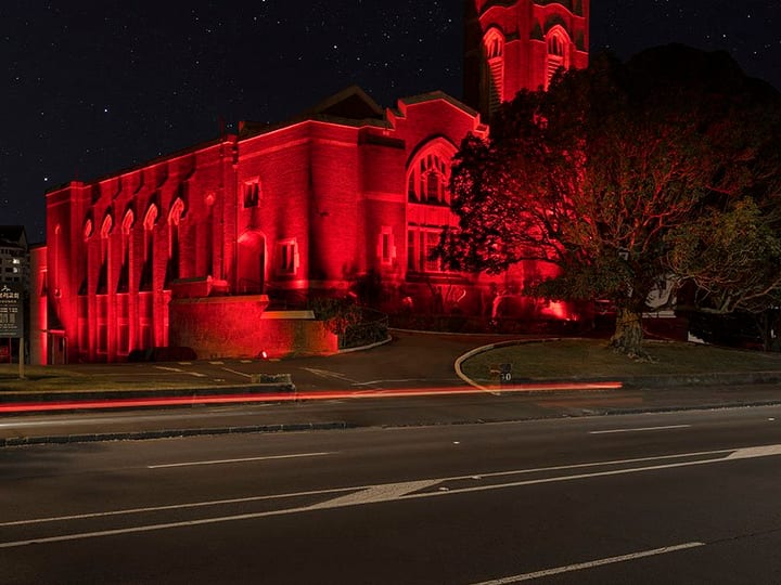 Red brick church with castellated tower, illuminated in red at night, viewed from across the road