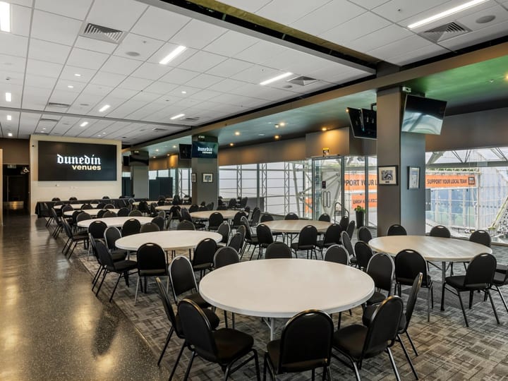 Stadium function room with round white tables, black chairs, wall-mounted screens, glass wall with field and stands beyond