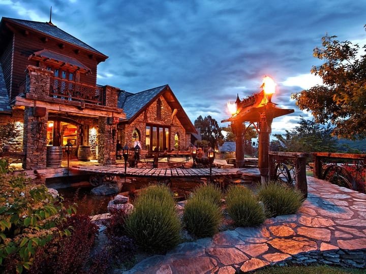 Stone and timber estate building at dusk with lit torch flares, pond, and garden terrace