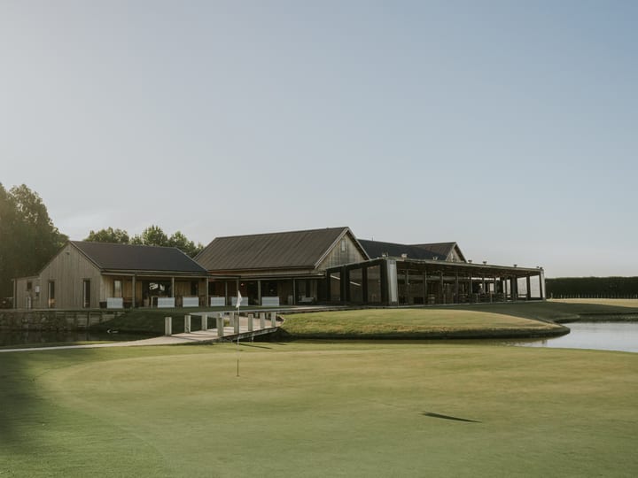 Timber and steel clubhouse beside a putting green with pond and trees, low afternoon light