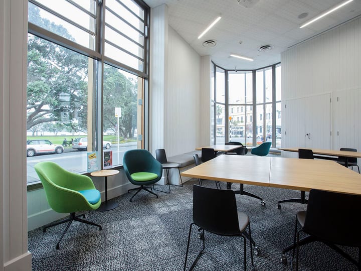 Curved-bay meeting room with floor-to-ceiling windows, green and teal chairs, light wood tables, street view