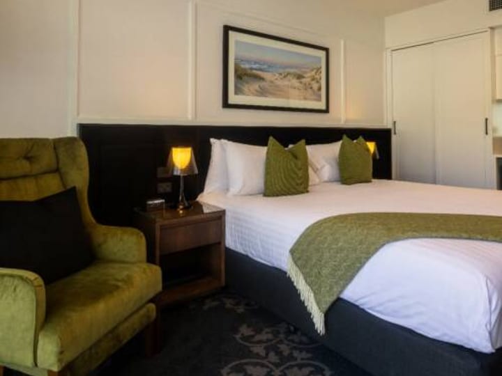 Hotel room with double bed, green velvet armchair, landscape artwork above headboard, warm lighting