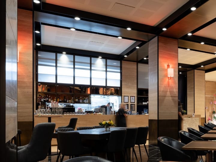 Dark-toned dining room with black leather chairs, round tables, wood-panelled walls, bar visible through opening