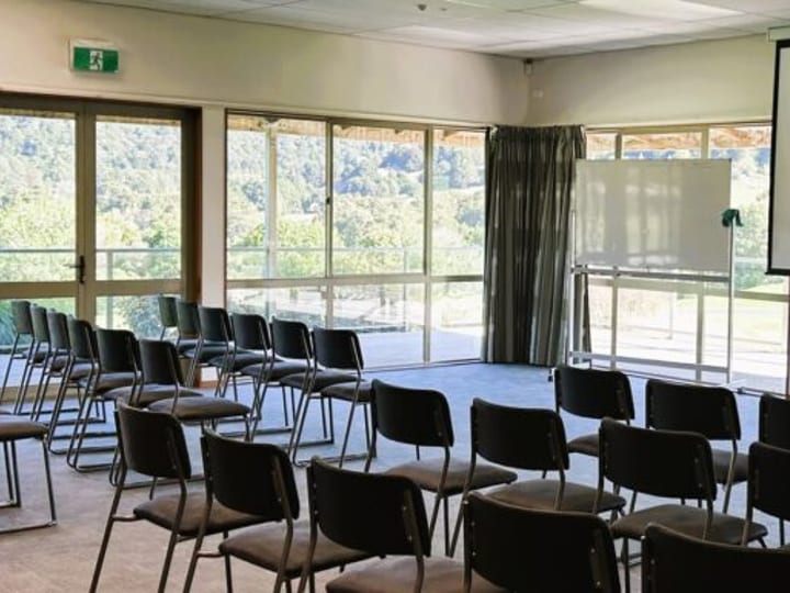 Meeting or function room with rows of chairs facing a projector screen, flanked by large windows overlooking greenery