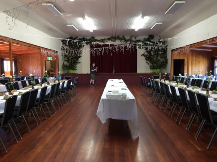 Timber-floored hall set with long white-draped tables, decorated with foliage and fairy lights, raised platform at far end