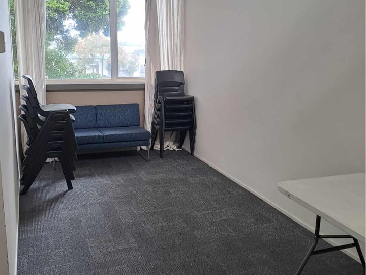 Small carpeted room with loveseat, stacked chairs, and table by window