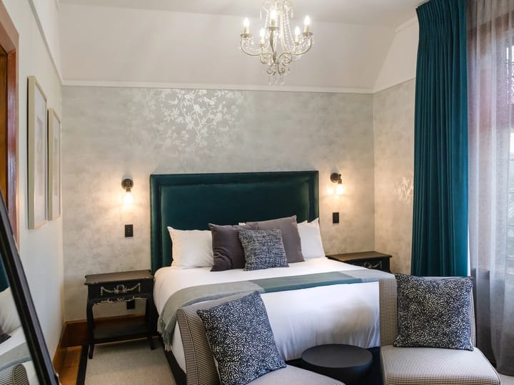 Guest room with dark green upholstered bed, crystal chandelier, patterned wallpaper, and stained-glass window