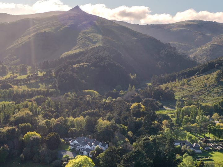Aerial view of a white manor house and gardens set in a valley with forested hills and a pointed peak