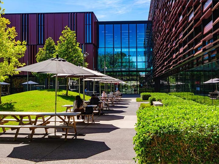 Campus courtyard with outdoor bench tables, umbrellas, and glass-clad buildings on either side