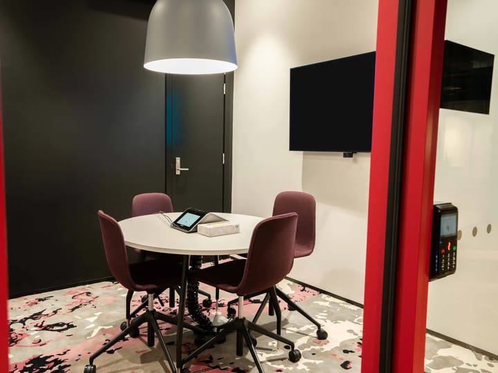 Small meeting room with round dark table, four upholstered chairs, whiteboard, wall-mounted screen, and patterned carpet