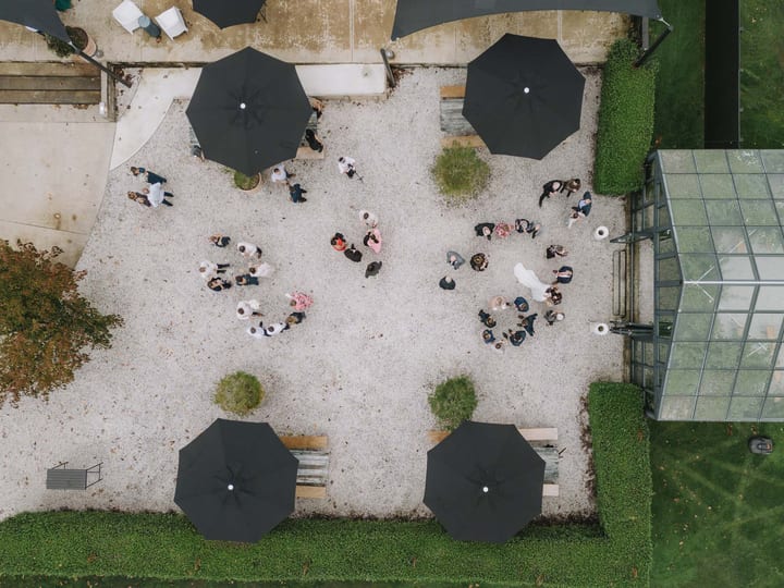 Aerial view of gravel courtyard with parasols and wedding guests mingling beside glass pavilion