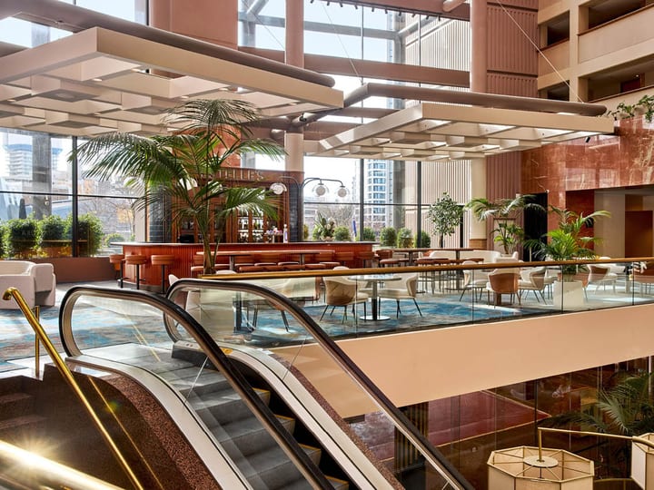 Multi-level atrium with escalators, indoor palms, pink stone walls, and a cafe bar below