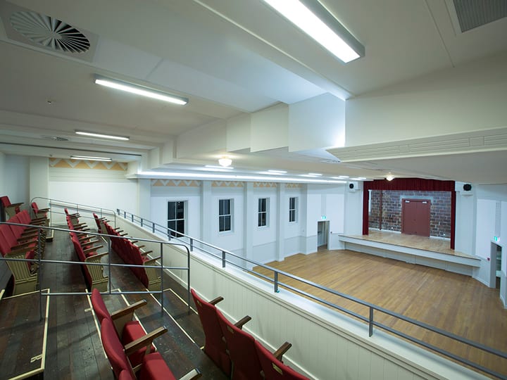 Hall interior with red theatre-style seating on upper balcony overlooking timber floor and stage