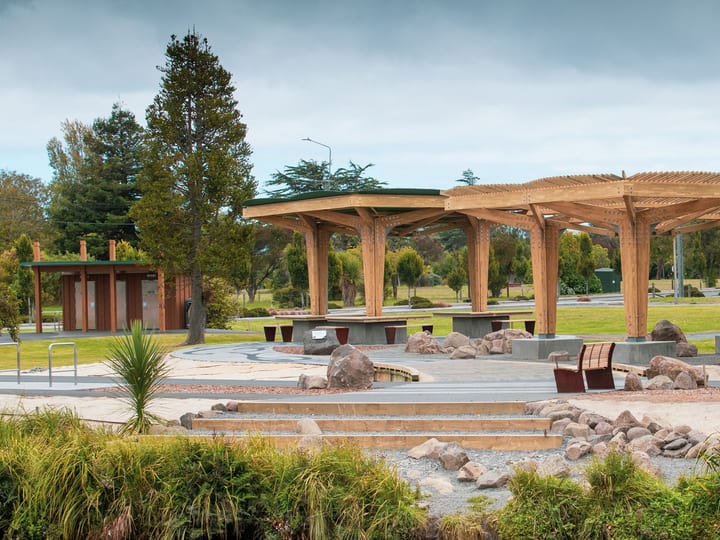 Timber open pavilion structure with latticed roof beams, concrete seating steps and boulders, parkland setting