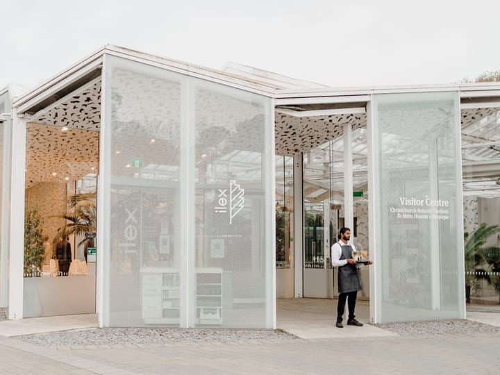White glazed pavilion exterior with perforated roof panels, timber columns and gravel surrounds