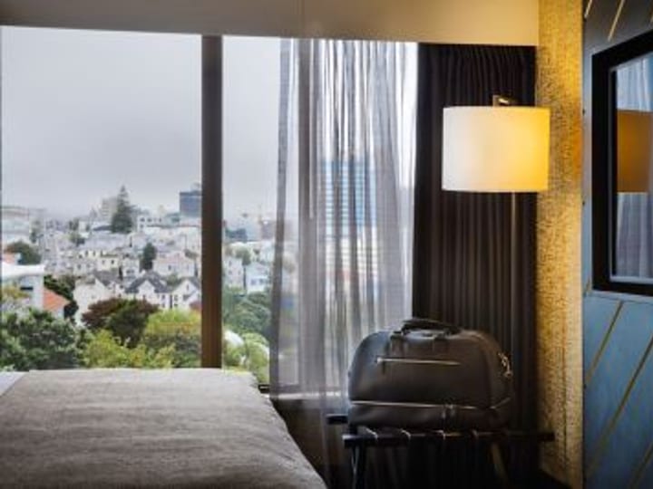 Hotel room with blue-teal walls, mustard pendant light, leather headboard, and city views