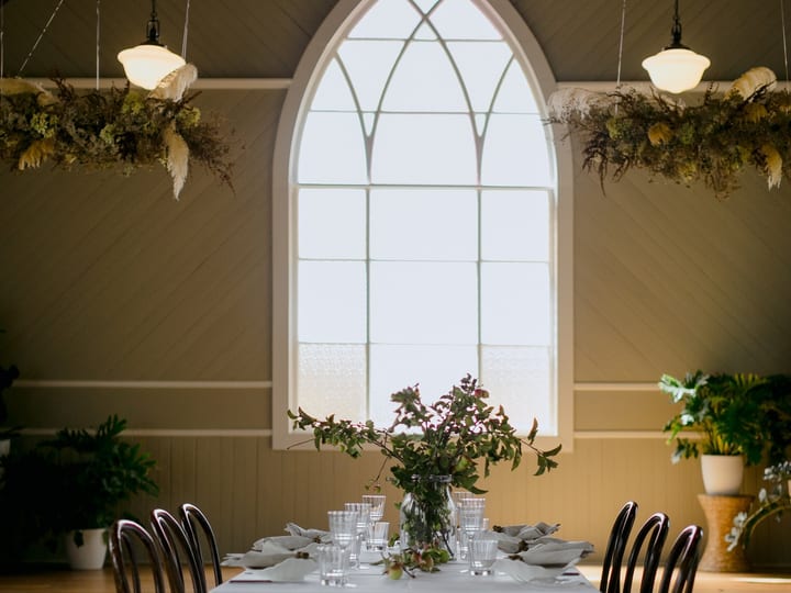 White-linen dining table beneath a Gothic arch window in a timber church interior with hanging floral arrangements