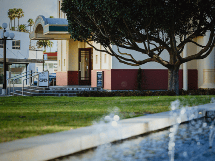 Art deco theatre exterior of cream and red render with a bare tree and water feature, street-level view