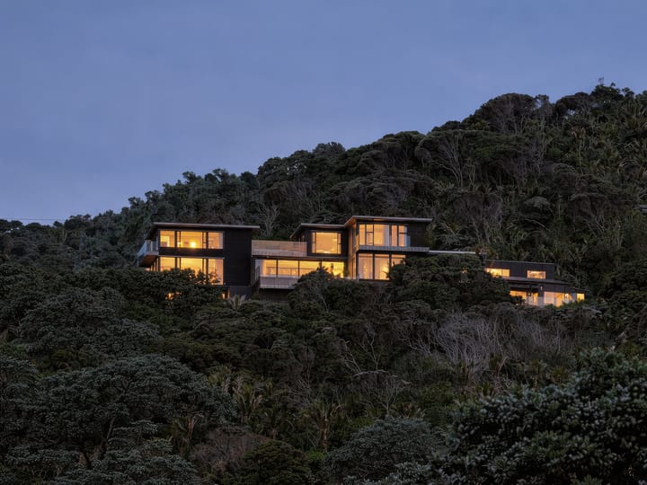 Dark timber buildings with illuminated windows on a steep bush-covered ridge at dusk