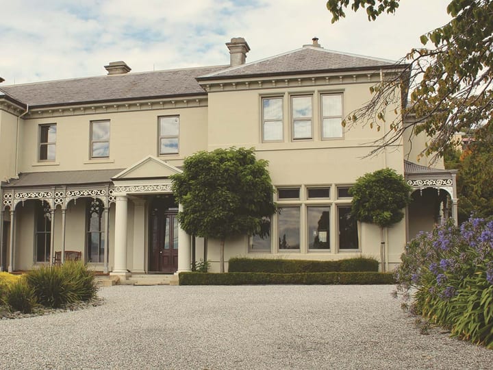 Two-storey cream Victorian manor with verandah and arched windows behind a gravel driveway and clipped hedges