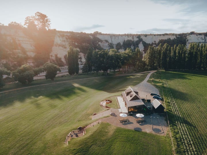 Aerial view of a small timber building set in flat green farmland beside white cliffs and a river bend