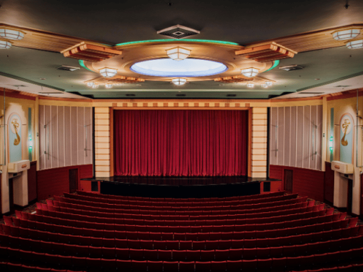 Art deco auditorium with tiered red seating, circular ceiling medallion, wall sconces, and a red curtained stage