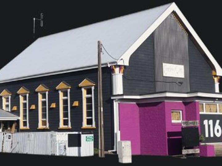 Dark-painted weatherboard building with pitched roof, purple brick addition, and yellow window trims