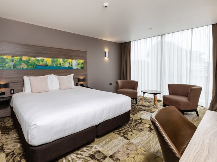 Hotel room with large bed, timber headboard, two armchairs by window with sheer curtains