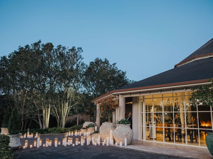 Low-lit exterior at dusk with rows of candles on a paved terrace, glazed pavilion and trees in background